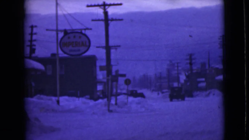 1946 Alaska frame with a car on a snow-covered road, prominent Imperial sign, utility poles, buildings, and distant hills.