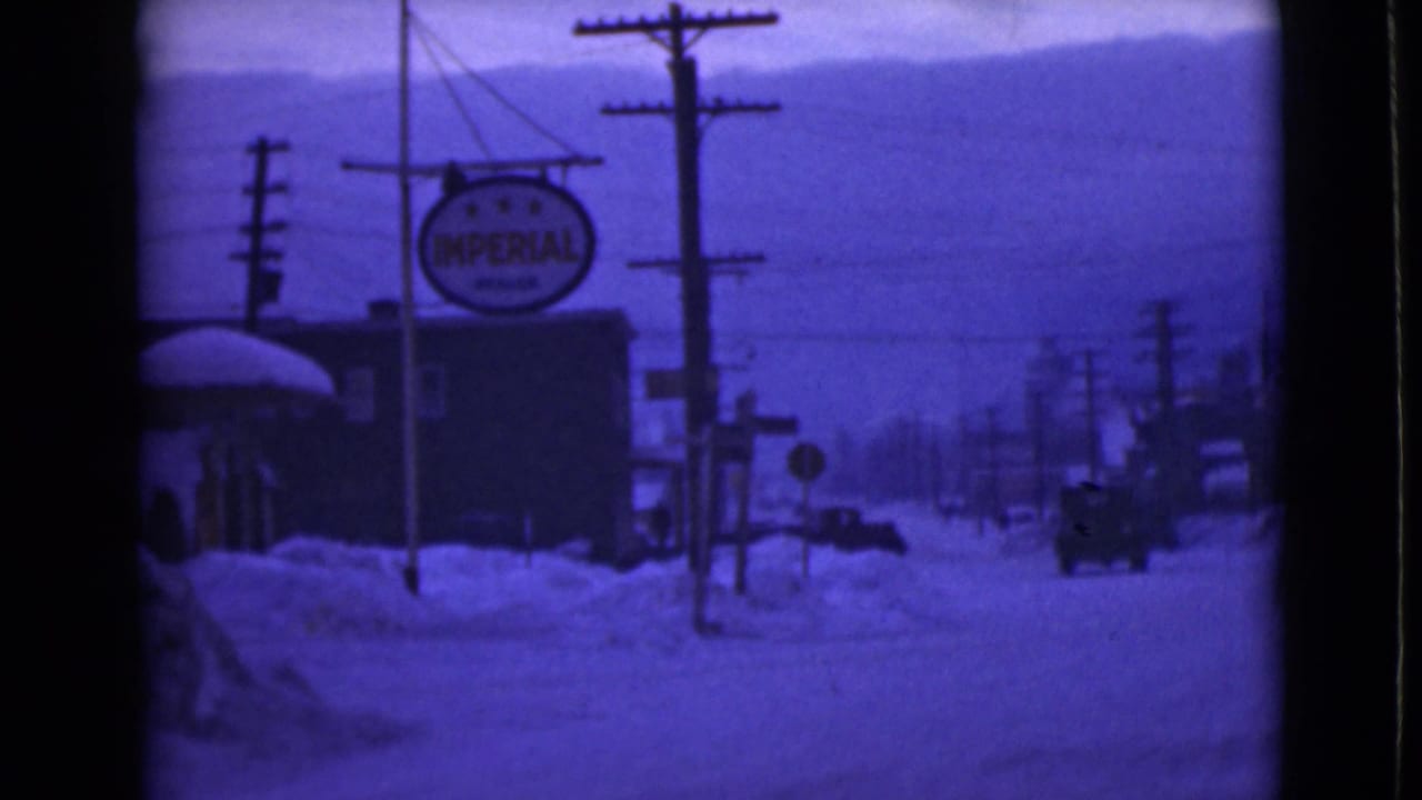 1946 Alaska home-movie frame showing a snowy street, deep drifts, utility poles, low buildings, and an Imperial Oil sign.