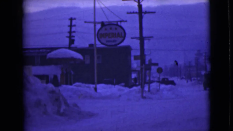 1946 Alaska street view with heavy snow, a hanging Imperial Oil sign, utility poles, buildings, and a distant vehicle.