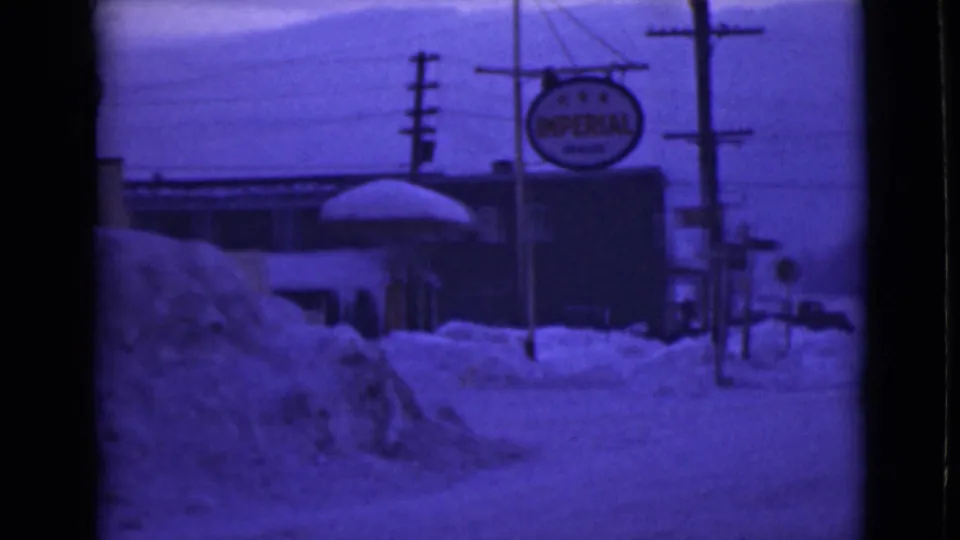 1946 Alaska frame showing deep roadside snow, an Imperial Oil sign, a snow-covered canopy, utility poles, and town buildings.