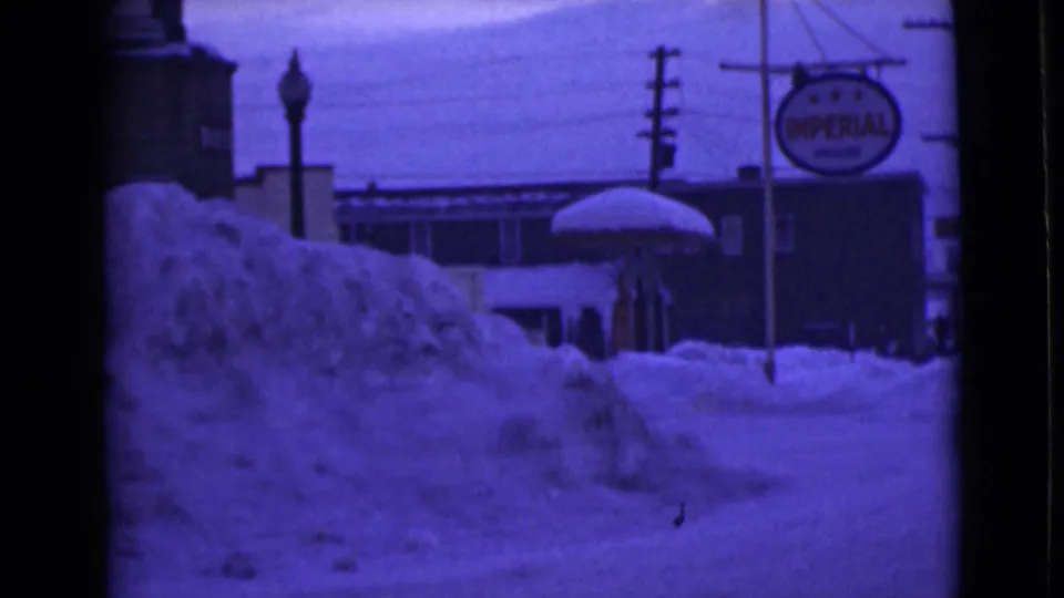 1946 Alaska home-movie still with high snowbanks, a roadside building, utility poles, and the Imperial Oil sign at frame right.
