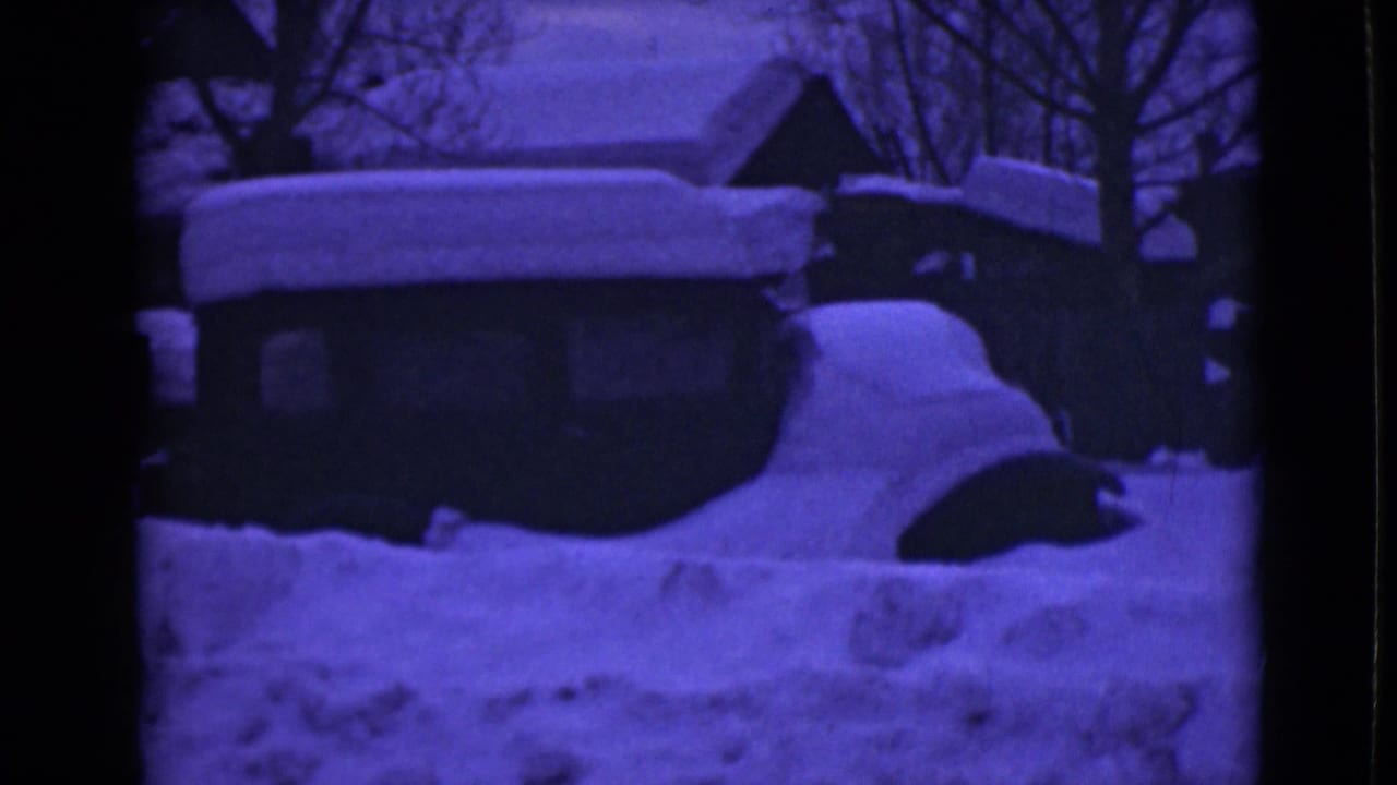 Deep snow covers a car and low buildings in a winter street scene.