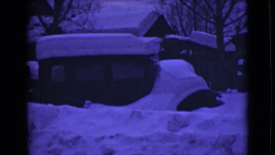 1946 Alaska home-movie frame showing heavy snowdrifts around a buried car, low buildings, rooflines, and bare trees.