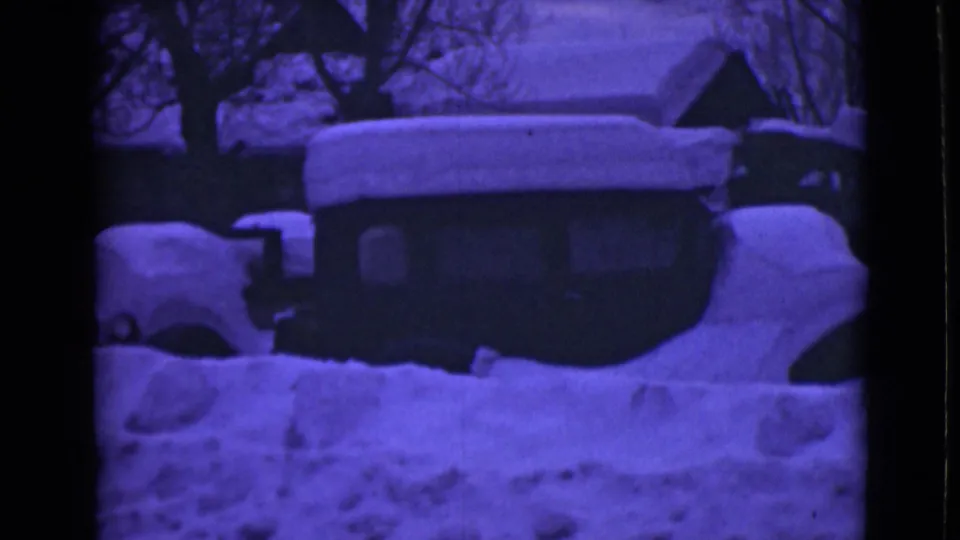 1946 Alaska winter frame with deep snow piled against a car and small buildings, tinted blue-purple by the 8mm film scan.