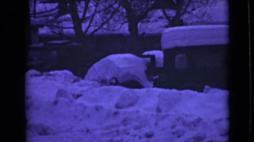 1946 Alaska frame showing a car nearly swallowed by snow, with a building edge, roofline, bare trees, and dense blue-purple film color.