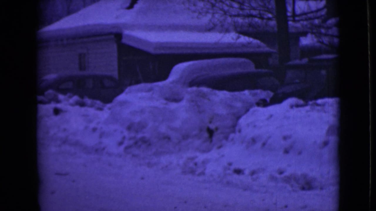 Snow-covered residential street with deep snowbanks, houses, bare trees, and parked cars.