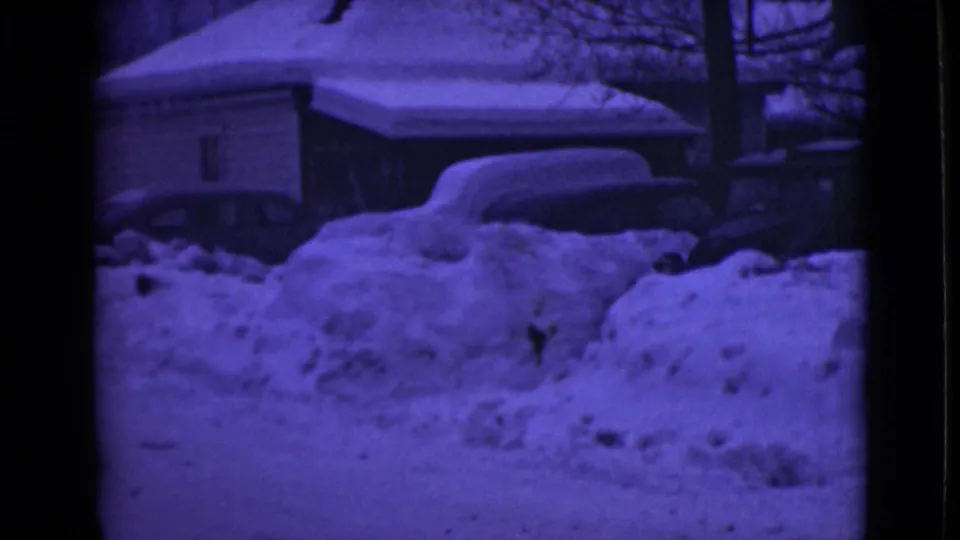 1946 Alaska home-movie frame showing a snowbound residential street with deep banks, houses, bare trees, and partially visible parked cars.