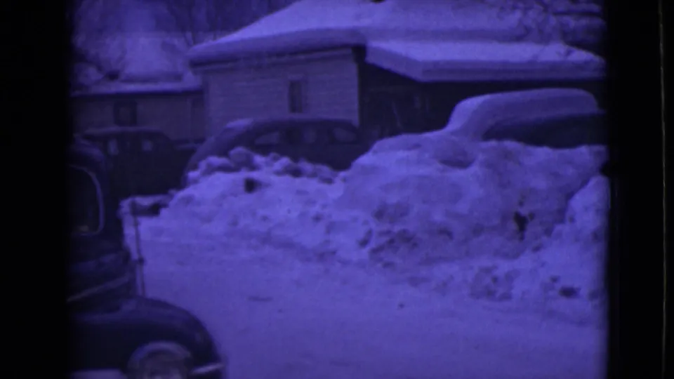1946 Alaska frame of a snowbank-lined residential street with houses in the background and a dark vintage car partly entering at left.