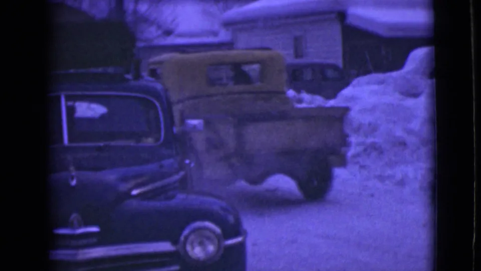 1946 Alaska home-movie frame with a dark sedan in front, a yellow truck moving behind it, and deep snow around nearby houses.