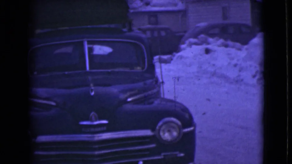 1946 Alaska frame showing a dark rounded sedan parked near deep snowbanks, with residential houses and winter trees behind it.