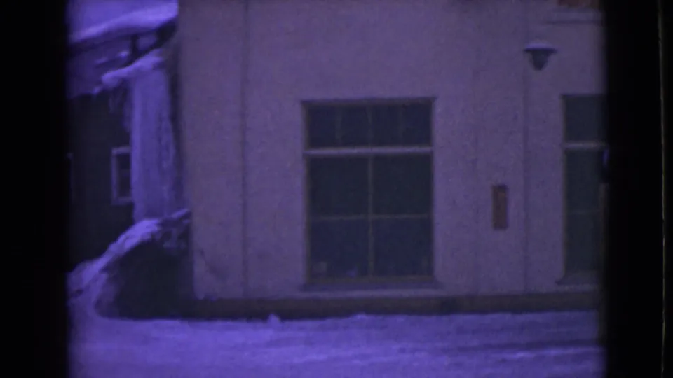 1946 Alaska home-movie frame with a pale building wall, broad window, snow at the base, visible icicles, and soft purple 8mm color.