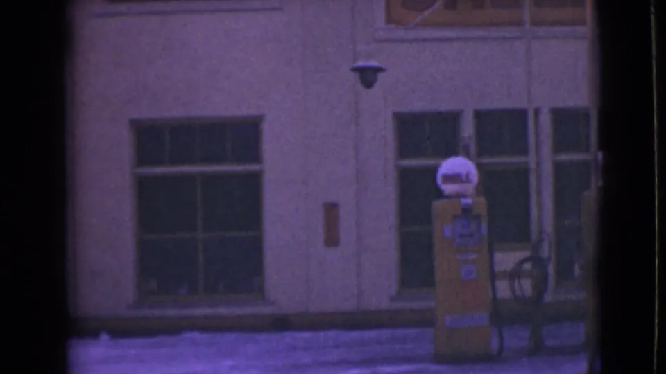 1946 Alaska home-movie frame showing a yellow Shell pump outside a snowy building with large windows and nearby dark metal furniture or equipment.