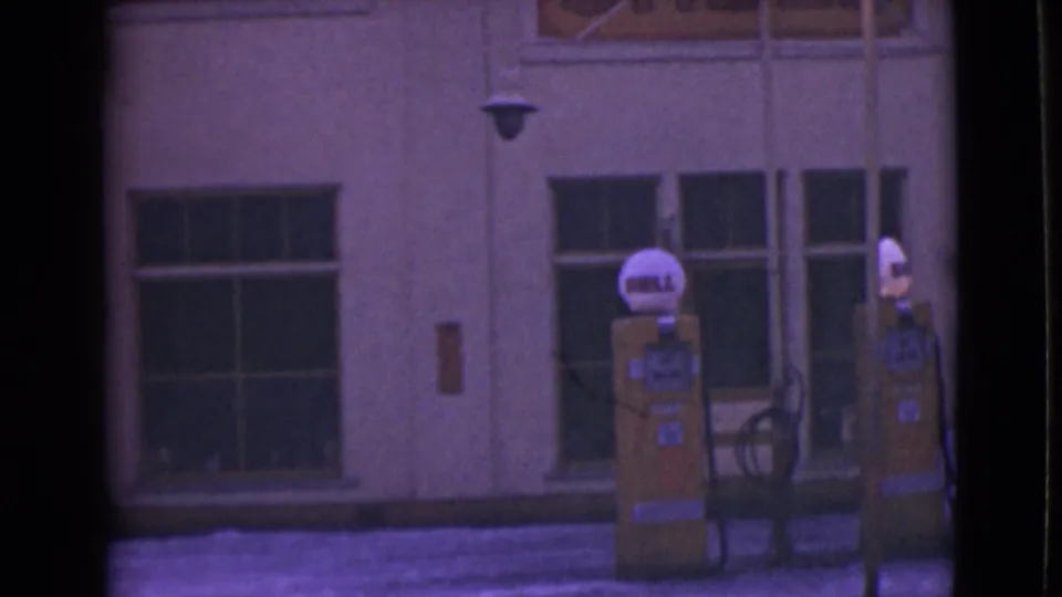 1946 Alaska 8mm frame showing two Shell gas pumps in front of a cream facade with dark windows and snow at street level.