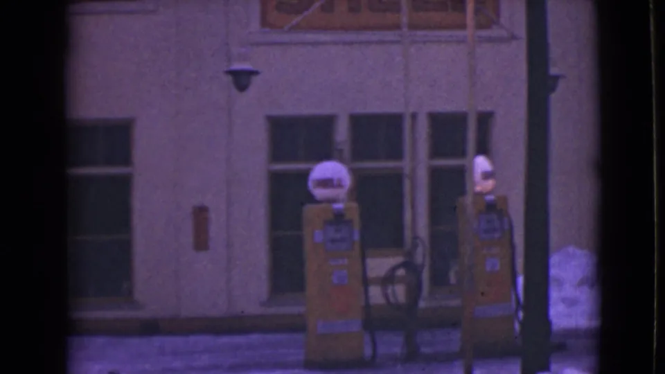 Soft 8mm frame of the Alaska gas station exterior, with Shell pump globes, yellow pumps, windows, and snow beside the curb.