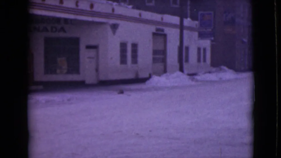 1940s 8mm view of a snow-covered roadside storefront frontage with Cranbrook B.C. Canada signage and winter snowbanks.
