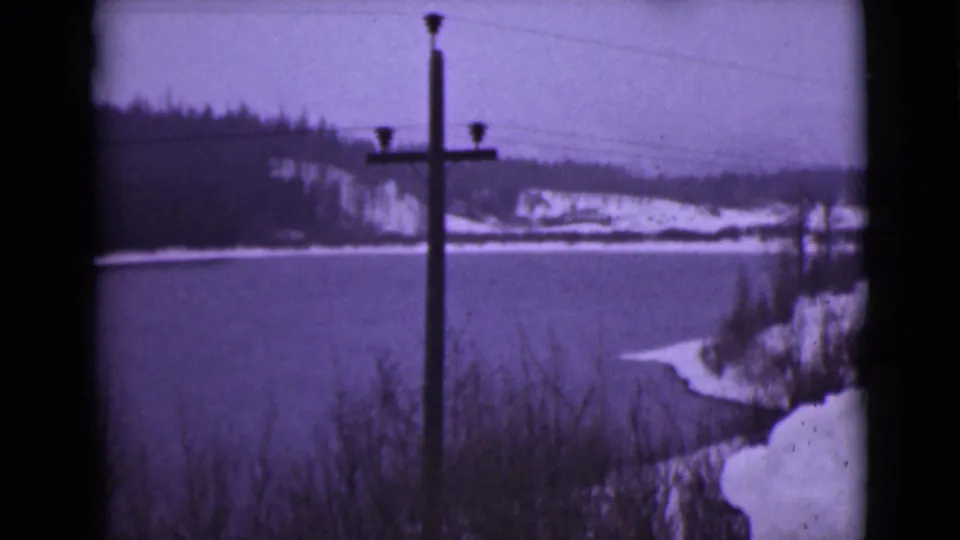 1946 Alaska 8mm frame of a winter lake scene, with shoreline brush, snowbanks, wooded hills, and a foreground utility pole.