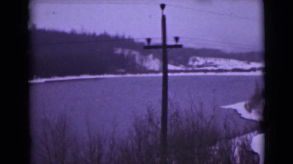1946 Alaska 8mm frame showing the same snow-edged lake and distant wooded hillside, with a utility pole silhouetted in the foreground.