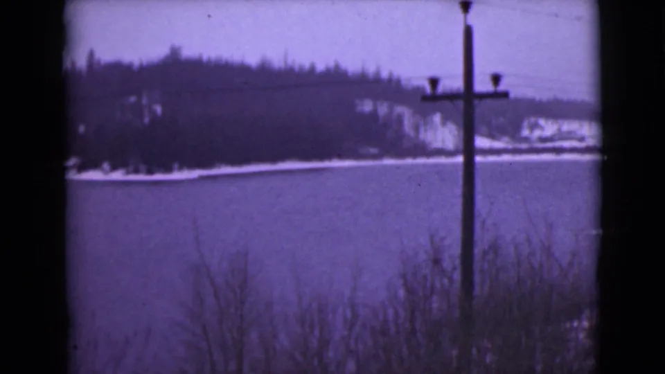 1946 Alaska home-movie frame with a still winter lake, distant wooded slope, shoreline snow, and a utility pole near the right side.