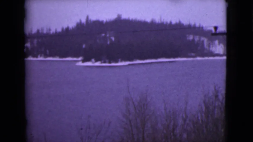 1946 Alaska home-movie frame of a winter lake and wooded hill, with overhead wires visible and foreground brush near the lower edge.