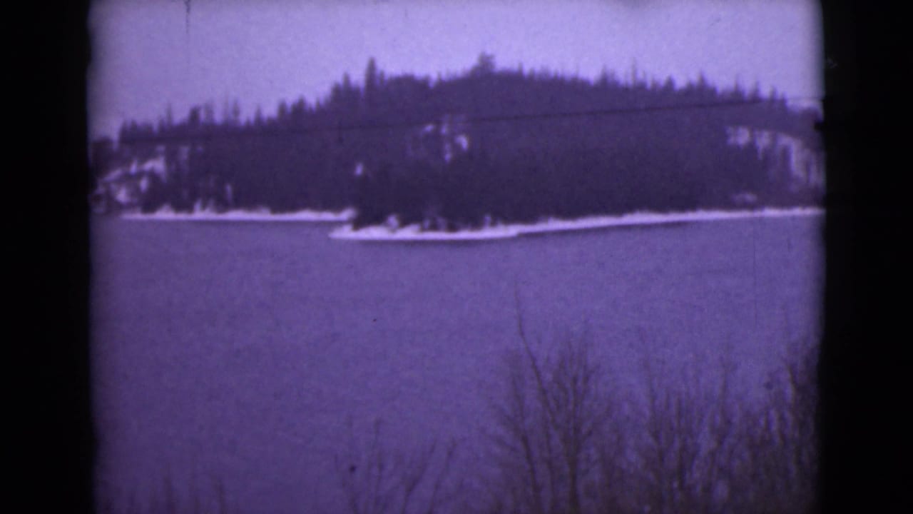 1946 Alaska 8mm frame showing a snow-edged wooded island or shoreline across frozen water, seen through a purple-tinted scan.
