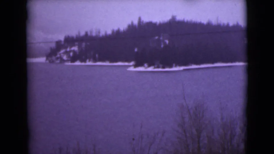 1946 Alaska home-movie frame of a dark evergreen island or shoreline bordered by snow across a frozen lake or inlet.