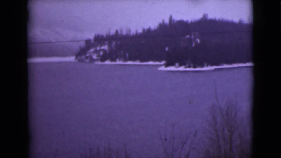 1946 Alaska 8mm frame with a snow-rimmed wooded shore and dark evergreens beyond a frozen expanse.