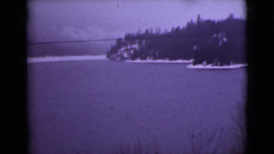 1946 Alaska home-movie frame showing a snow-edged wooded point across frozen water, with faint hills in the background.
