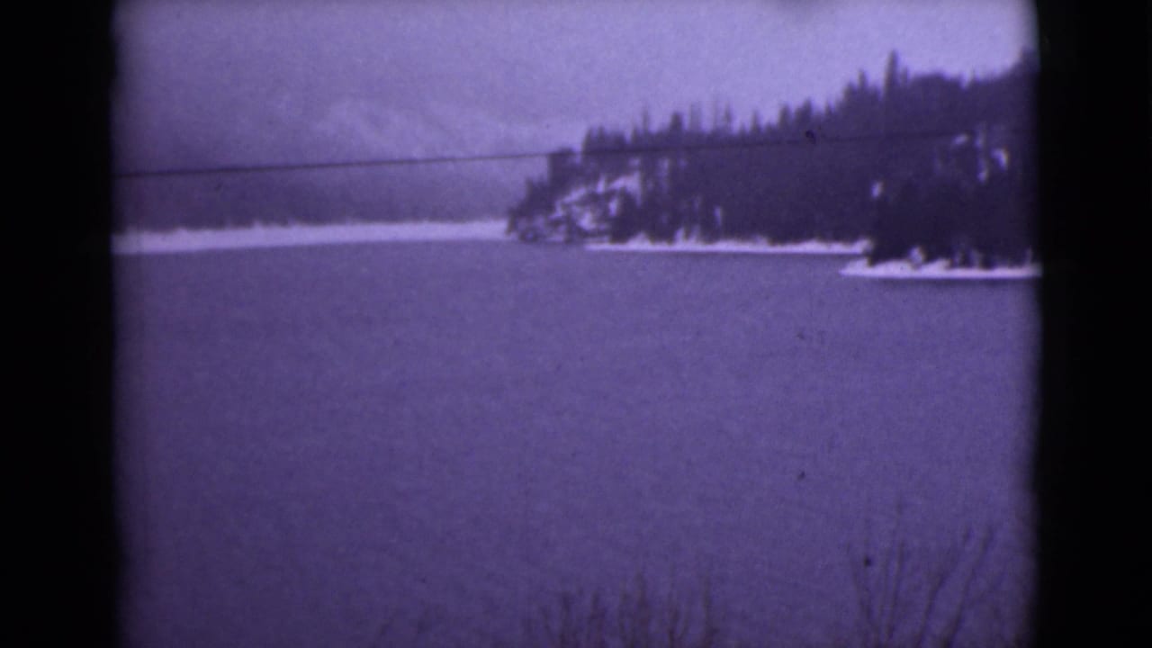 Snowy lake with evergreen shoreline and distant mountains under a purple-tinted sky.