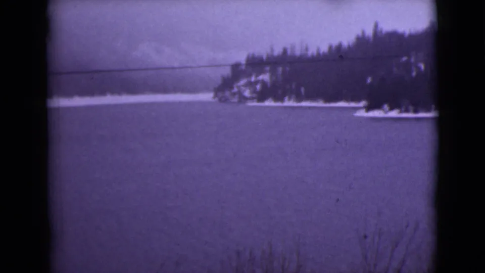 1946 Alaska 8mm frame showing a broad snowy lake, dark evergreen shoreline, and distant mountain slopes with visible film gate edges.