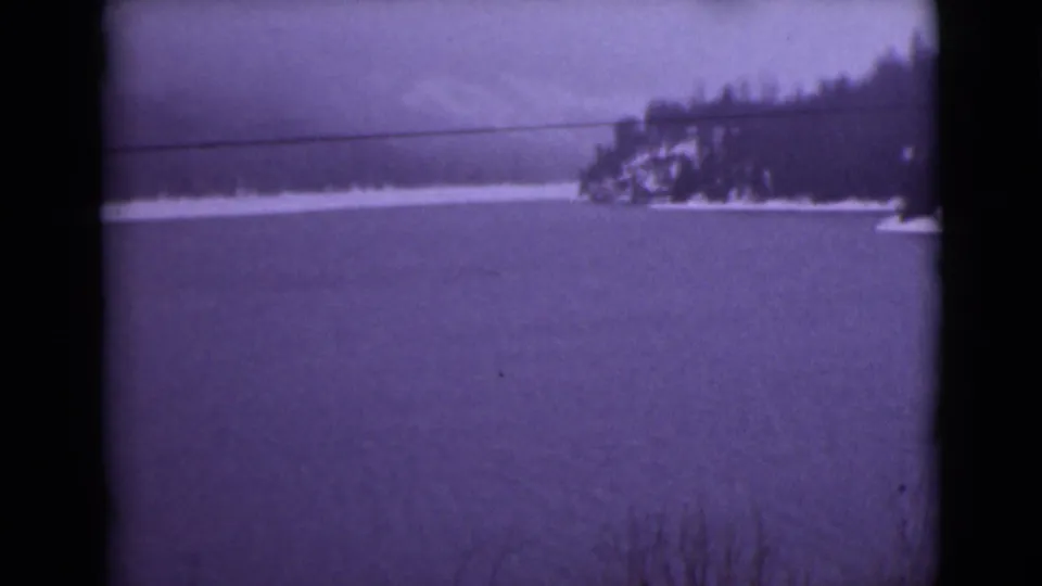 1946 Alaska home-movie frame showing a snow-covered lake, wooded shoreline point, and distant mountain ridge through soft 8mm color.