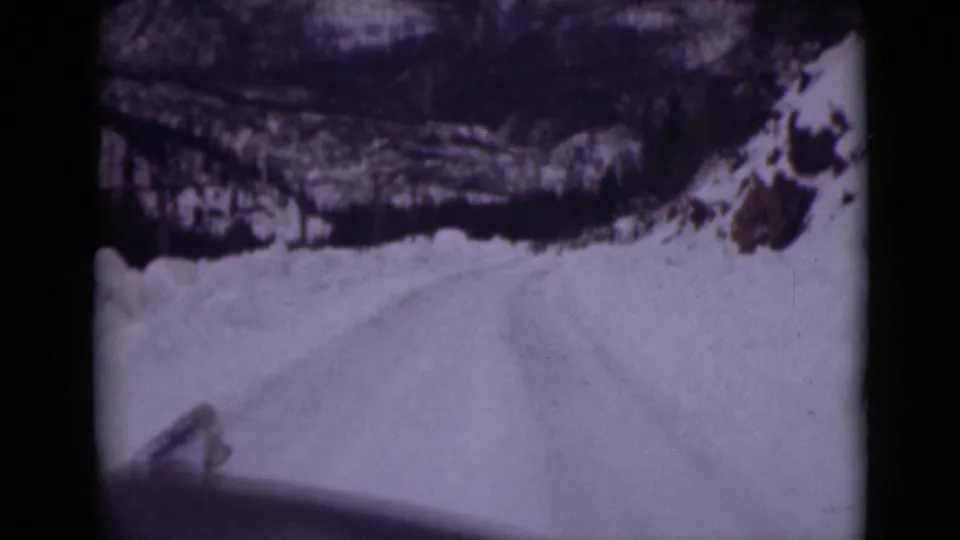 1946 Alaska 8mm travel frame looking ahead along a snow-covered road through rocky banks and dark winter forest from a vehicle viewpoint.