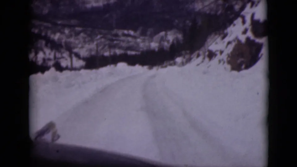 1946 Alaska home-movie frame showing a snow-covered mountain road, rocky roadside, dark trees, and part of the vehicle at the bottom edge.