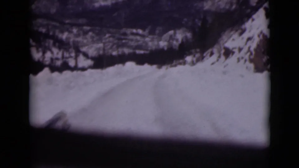 1946 Alaska home-movie frame showing a snowy road or slope through dark car-window framing, with rocks and snowbanks ahead.