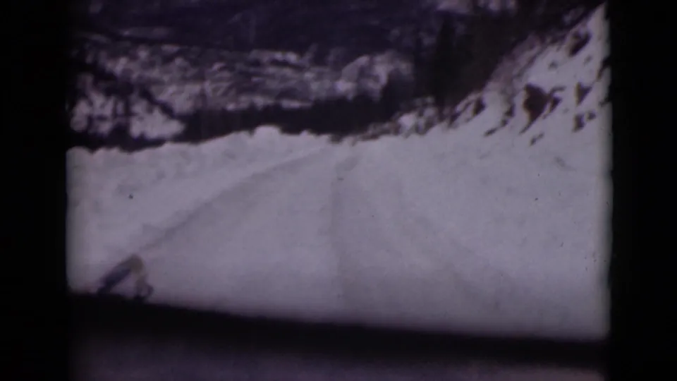 Snow-covered road or pass in Alaska, filmed through a car window with dark frame edges and rocky terrain above the snow.