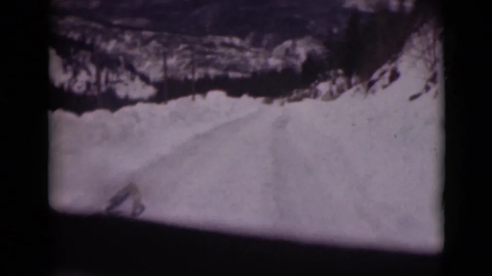A soft 8mm frame of snowy Alaska road travel, with a dark car-window border and a pale road surface below a rocky slope.