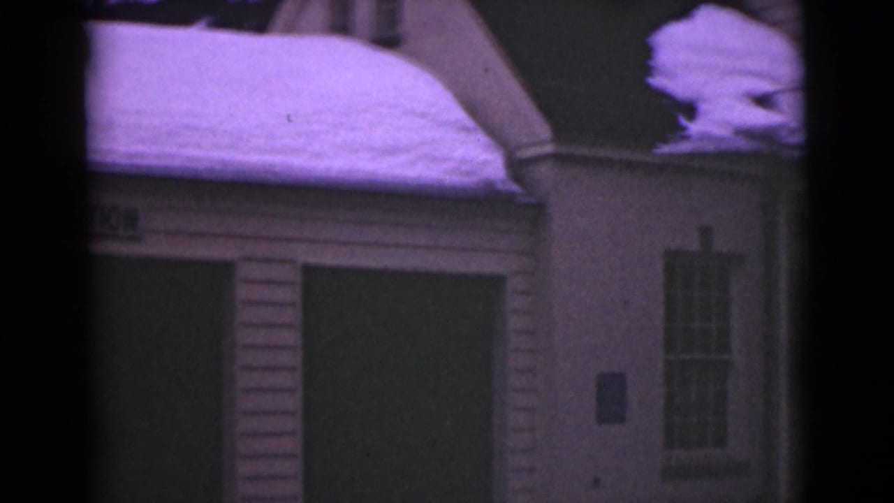 Snow-covered clapboard building with dark doorway, window, and heavy roof snow.