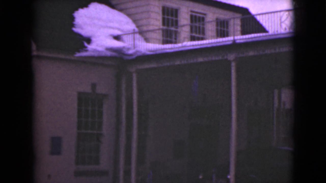 1946 Alaska 8mm frame showing a wooden building facade with upper windows, a porch edge, snow along the roof, and balcony railing.