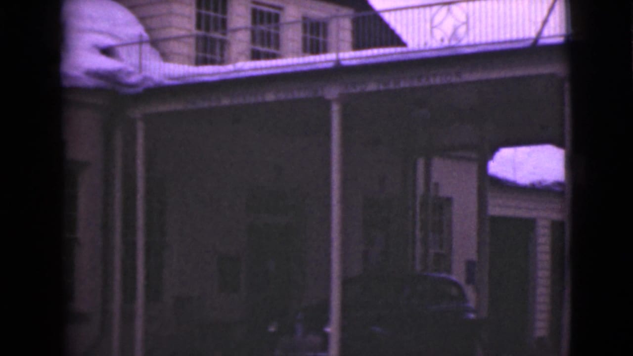 Building exterior with upper windows, porch columns, railing, snow, and a parked car partly visible.