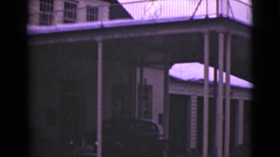 1946 Alaska home-movie frame showing the porch corner, vertical columns, nearby snow-covered roof, windows, and a dark car below.