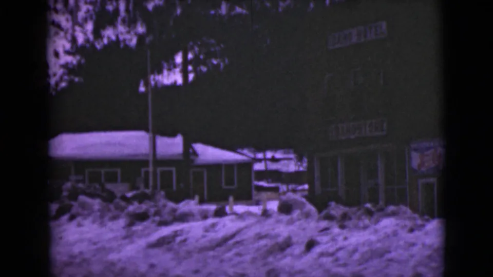 1946 Alaska 8mm frame with piled snow before storefront doors, a low roofline, and partially visible hotel signage.