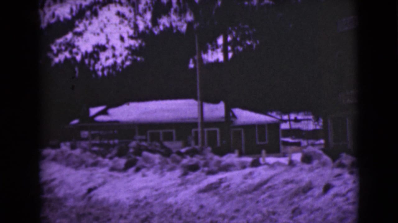 Snow-covered house with high snowbanks, trees, and a flagpole in a purple-tinted 8mm frame.