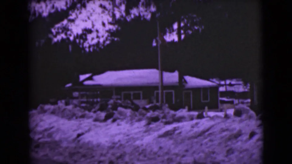 1946 Alaska winter exterior with a house behind piled snow, framed by trees and a nearby flagpole.