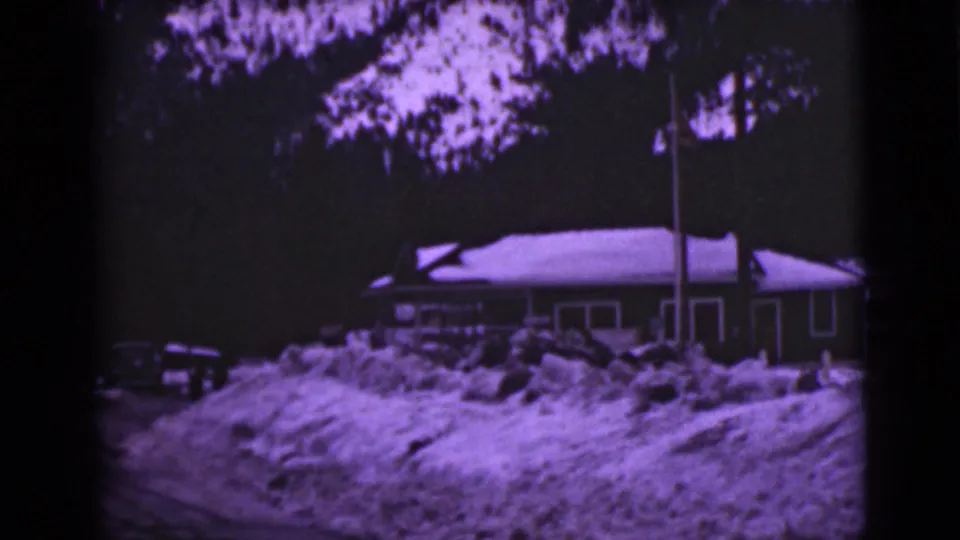 1946 Alaska home-movie frame showing a quiet winter house exterior with piled snow, trees, and a flagpole.