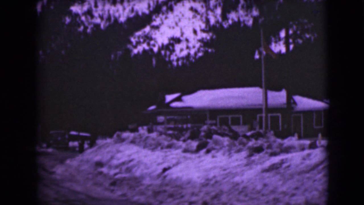 Snow-covered house beside a snowy road with a parked car and flagpole.