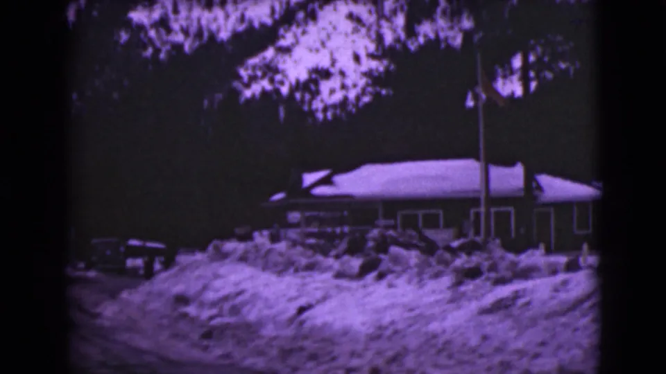 1946 Alaska 8mm frame showing a snow-covered house, deep roadside drifts, a parked car, trees, and a flagpole.