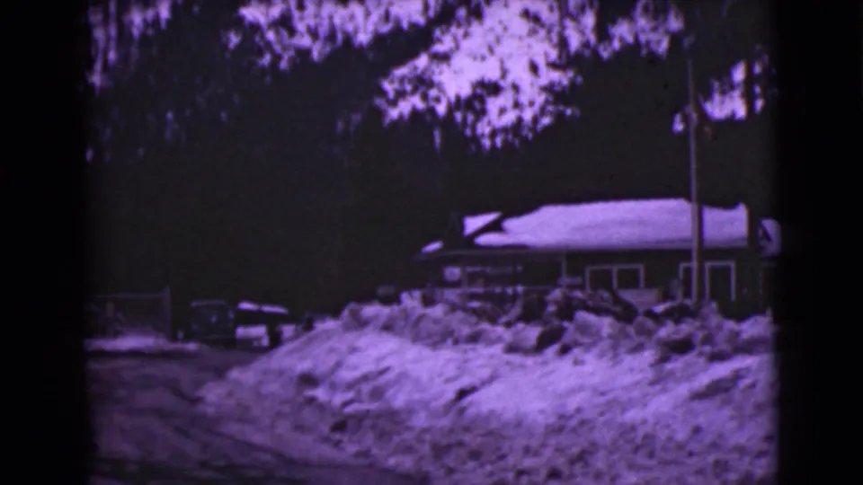 1946 Alaska home-movie frame of a low building under snow, with a parked car, flagpole, trees, and heavy drifts.