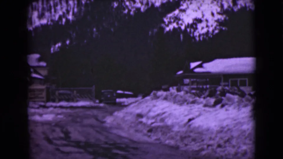 1946 Alaska home-movie frame showing a snow-covered road, large drifts, a parked car, a house edge, and dark trees.