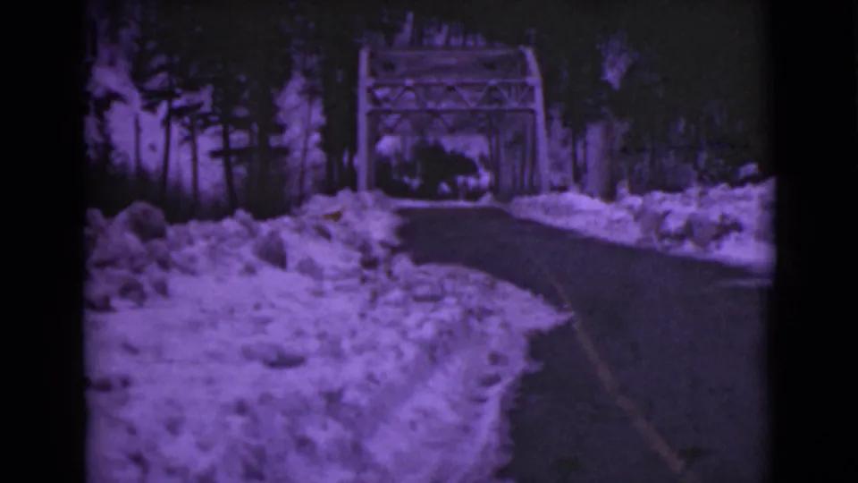 1946 Alaska 8mm frame from a moving road view, with snowbanks and a metal truss bridge ahead among dark trees.