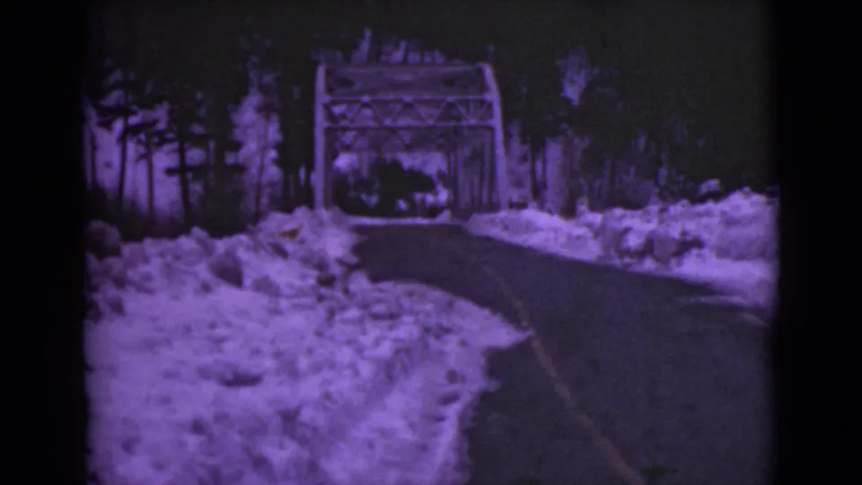 1946 Alaska 8mm frame showing the road near a metal truss bridge, bordered by snowbanks and evergreen forest.