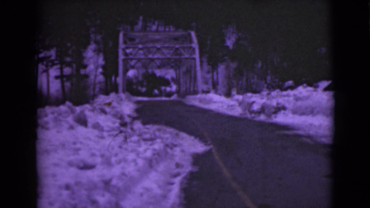 1946 Alaska 8mm frame showing a cleared road bordered by deep snowbanks and trees as it approaches a metal truss bridge.